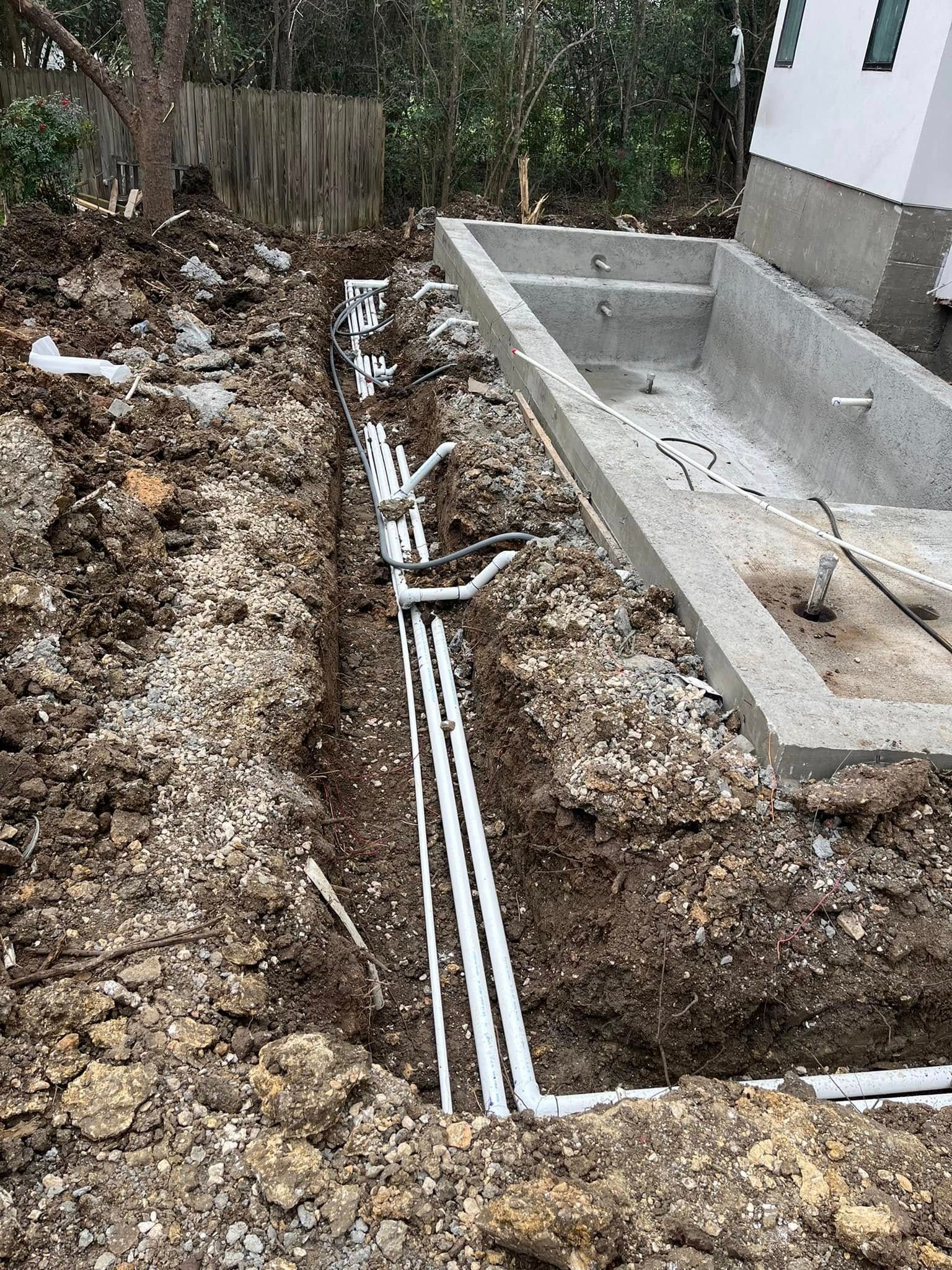 Outdoor trench with white PVC pipes beside a concrete foundation and yard debris.