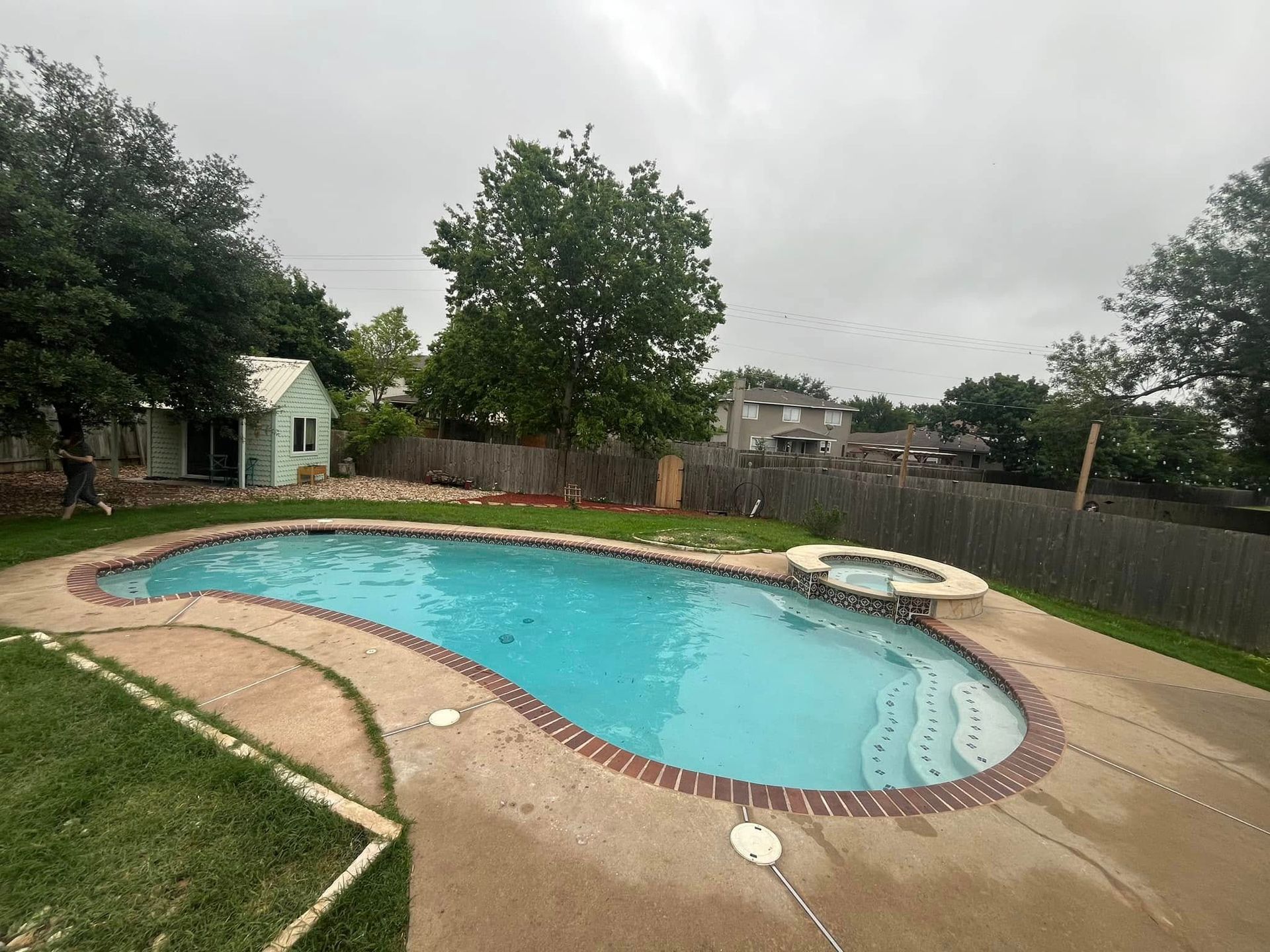 Backyard swimming pool with a curved concrete deck, fenced yard, and cloudy sky