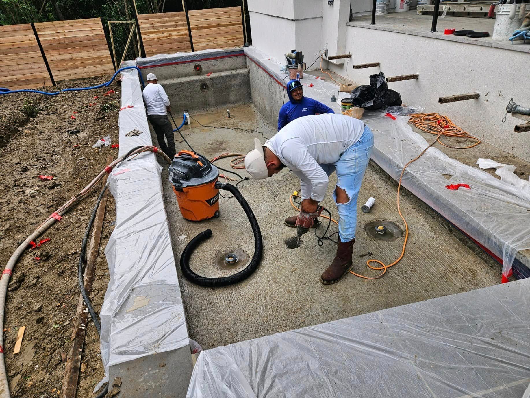 Workers installing concrete in a rectangular backyard foundation pit with a vacuum and hoses.