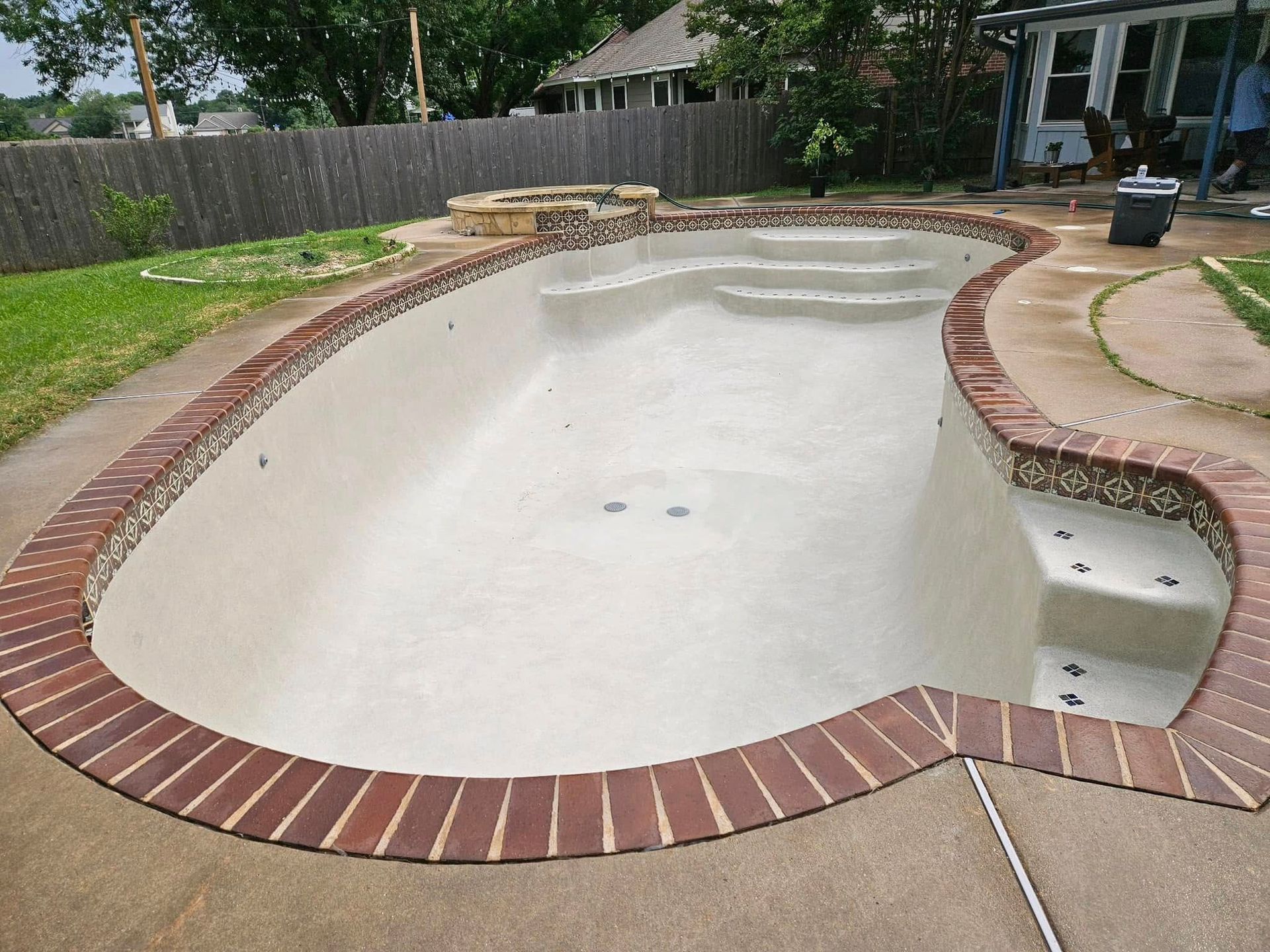 Empty backyard swimming pool with white interior and brick coping, surrounded by grass and patio.