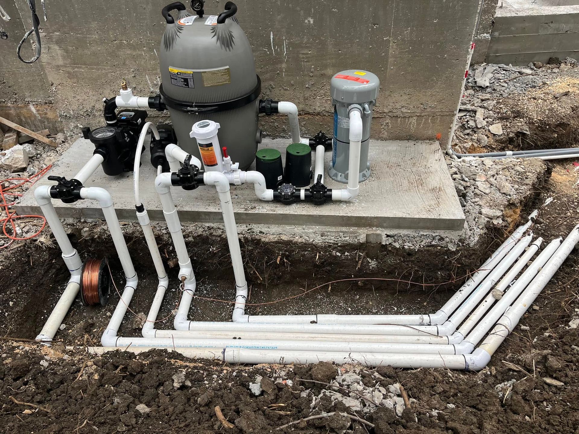 Outdoor pool plumbing and filter equipment with white PVC pipes and pumps beside a concrete wall.