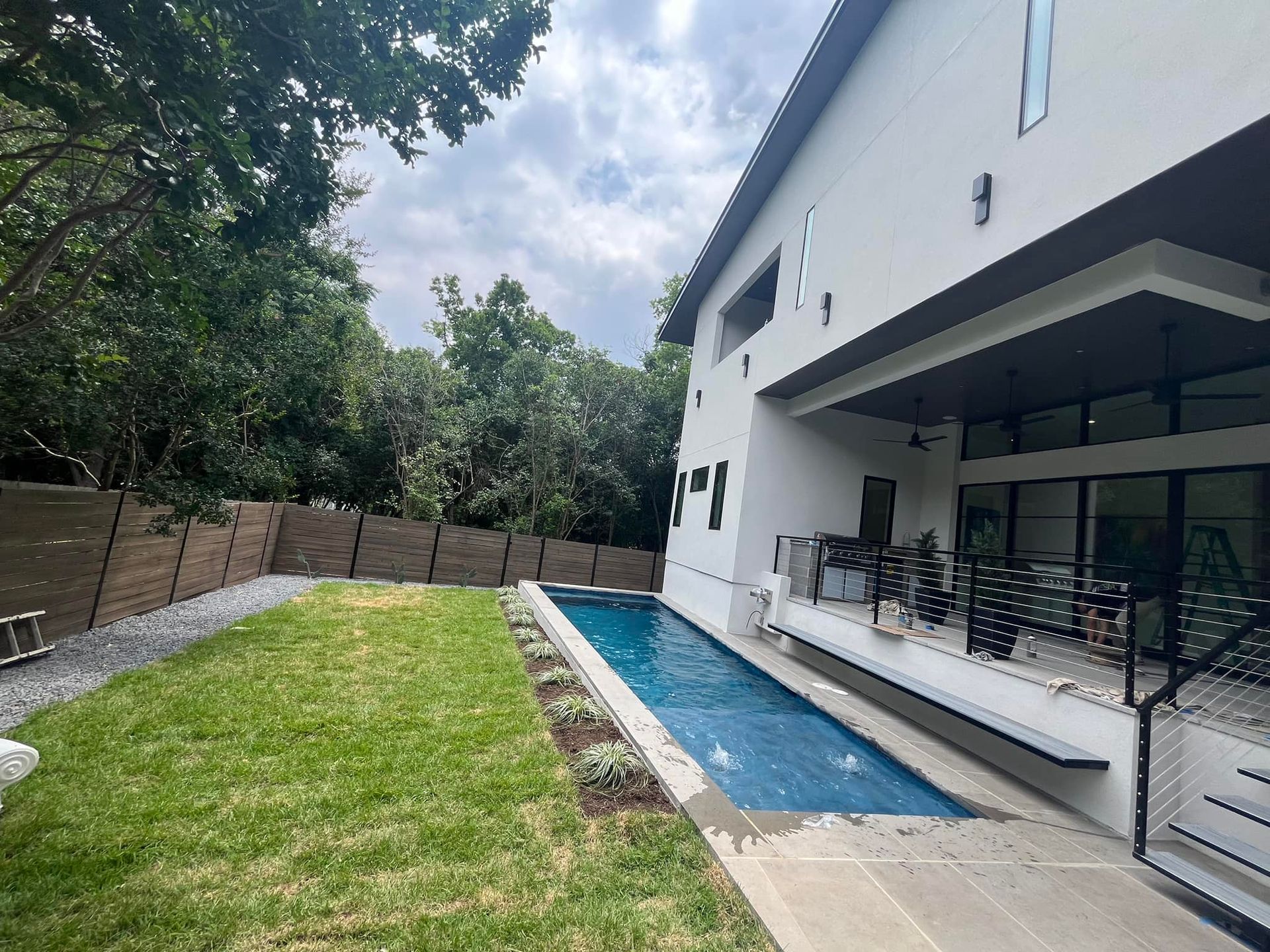 Backyard pool beside a white house with a grassy lawn and trees along the fence
