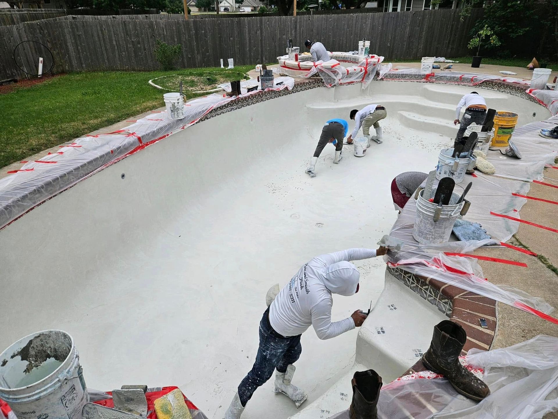 Workers resurfacing an empty swimming pool with white coating and red tape around the edge