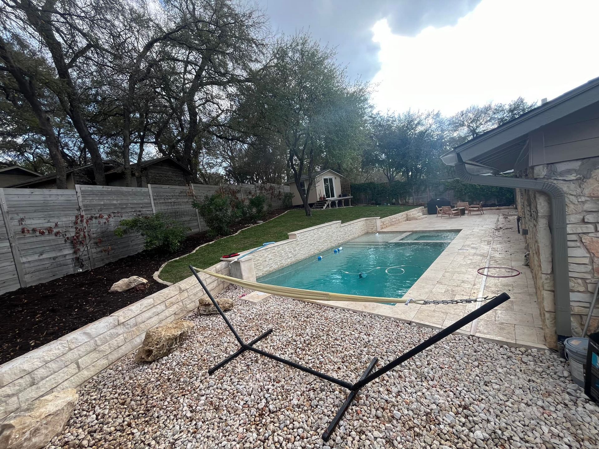 Backyard swimming pool with stone deck, lounge chairs, and trees under a cloudy sky