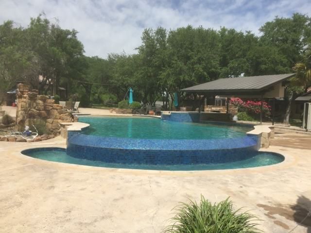 Outdoor resort pool with a curved blue swim-up edge, lounge chairs, and trees under a cloudy sky