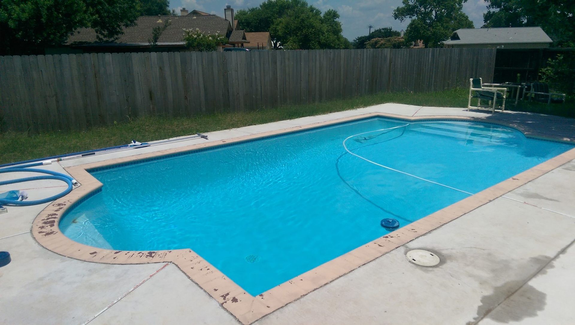 Backyard swimming pool with bright blue water, concrete deck, wooden fence, and patio furniture