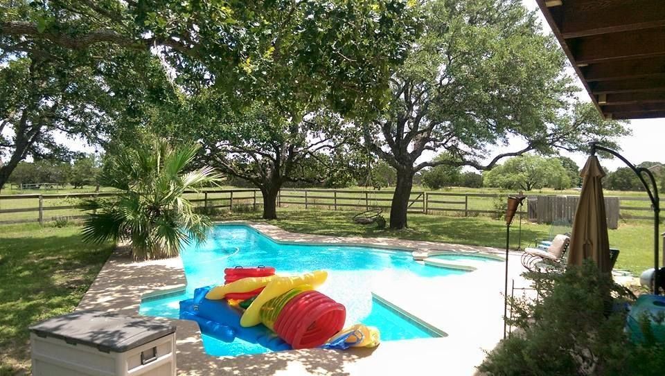 Backyard pool with colorful inflatables, lounge chair, and large trees beyond a wooden fence