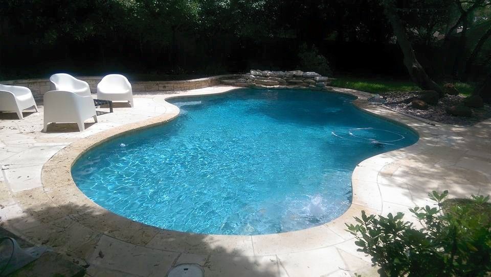 Curved blue swimming pool surrounded by a light stone deck and white lounge chairs in a shaded backyard.