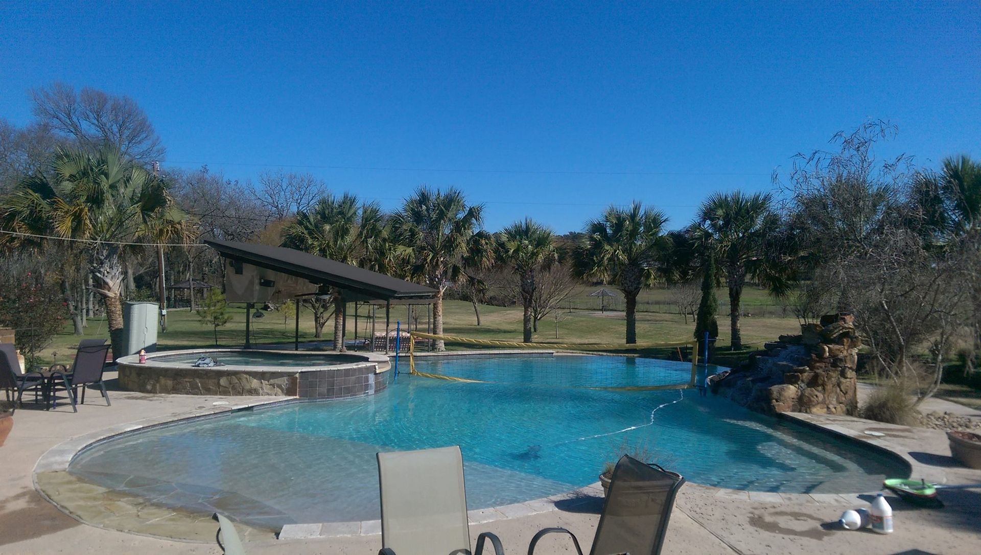 Backyard pool with lounge chairs, covered seating area, and trees under a clear blue sky
