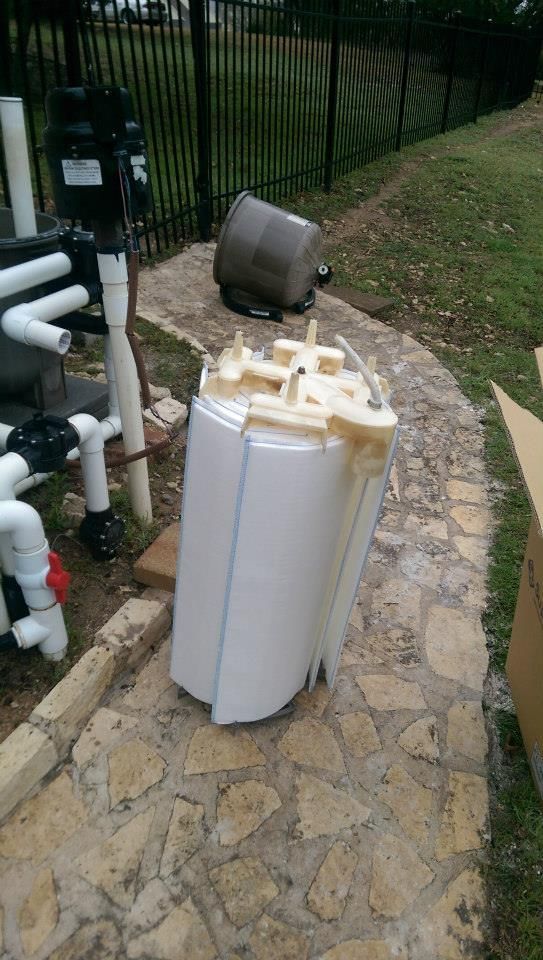 Outdoor pool equipment with white filter tank, pipes, and pump on a stone walkway near a fence.
