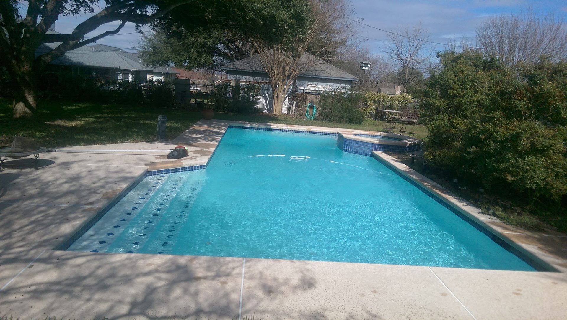 Backyard swimming pool with turquoise water, surrounded by trees and patio seating in bright daylight