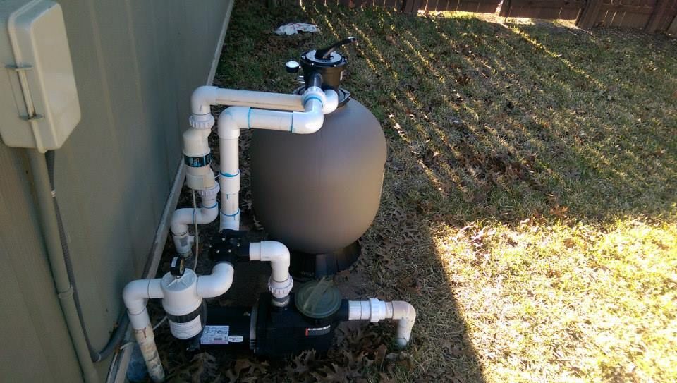 Outdoor pool pump and filtration equipment beside a house, with pipes, valves, and a tan tank.