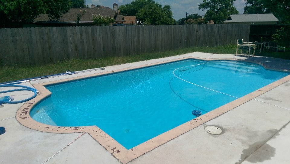 Empty backyard swimming pool with bright blue water, concrete deck, and wooden fence.