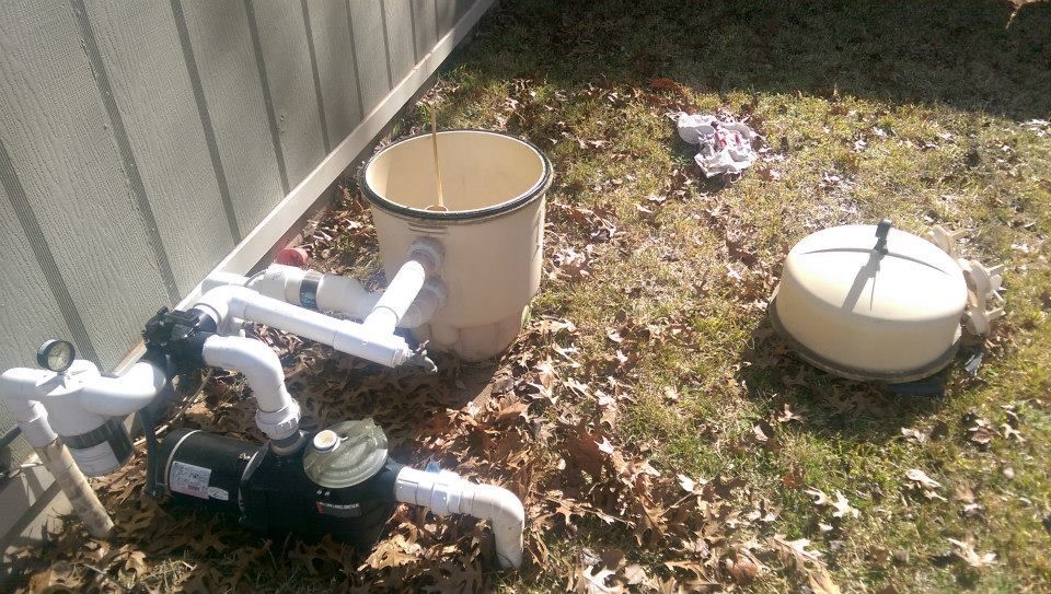 Outdoor pool equipment and plumbing beside a fence, with two beige filter tanks on grass and leaves.