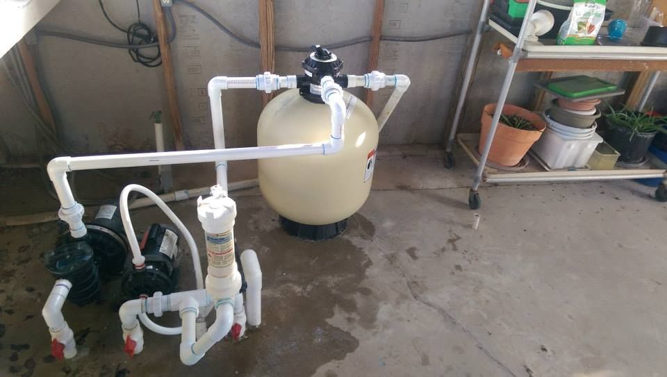 White plumbing pipes and a pump connected to a round beige tank in a concrete utility area.
