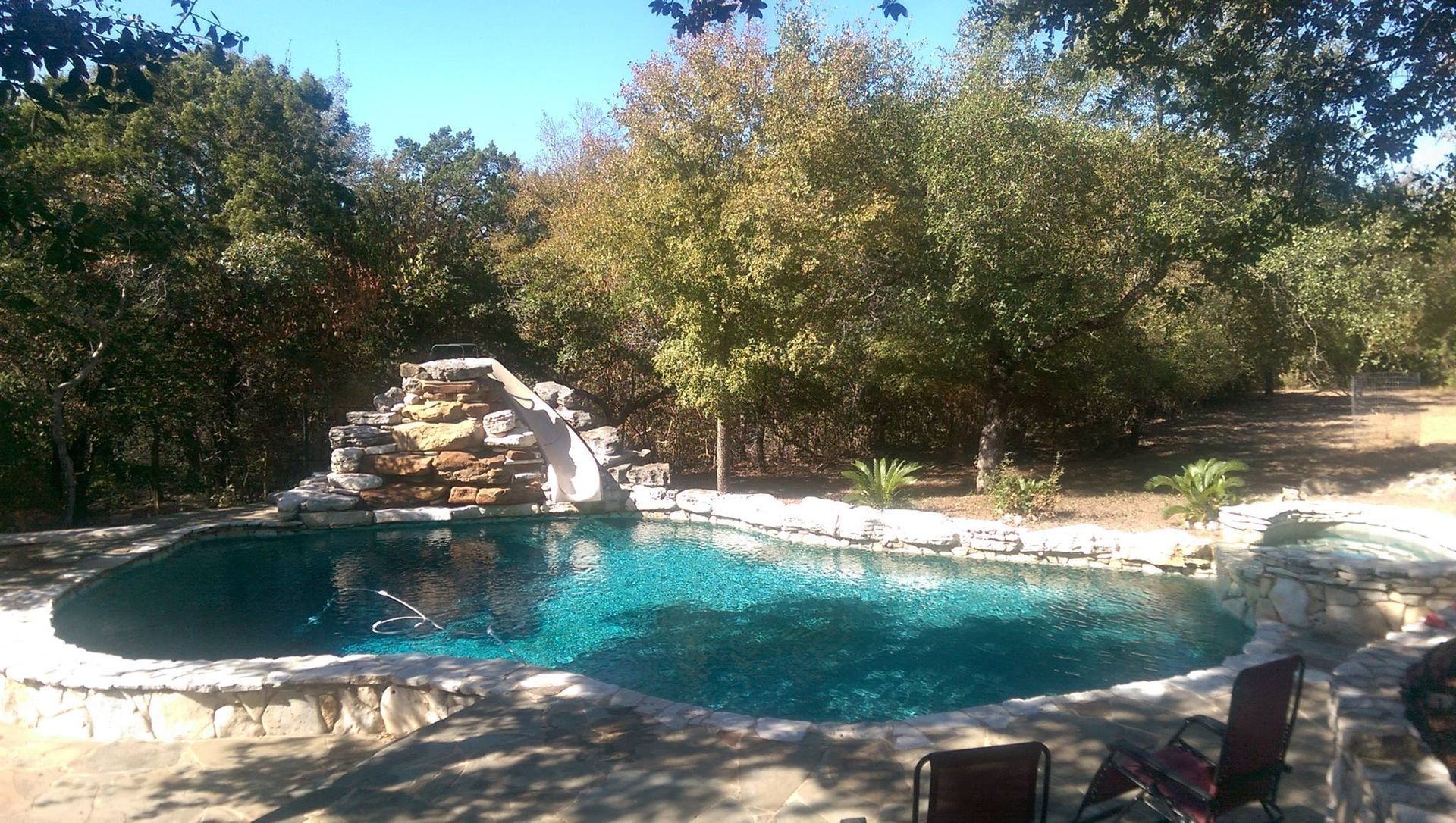 Backyard pool with a rock waterfall, surrounded by trees and lounge chairs under sunny skies