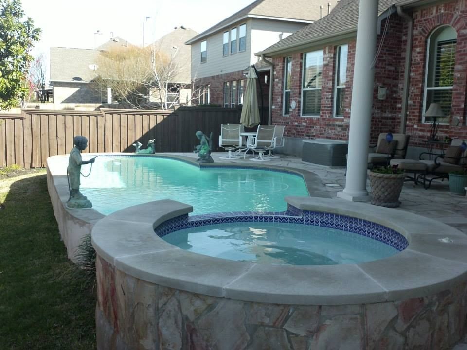 Backyard pool and spa beside a brick house, with patio chairs and a wooden fence.