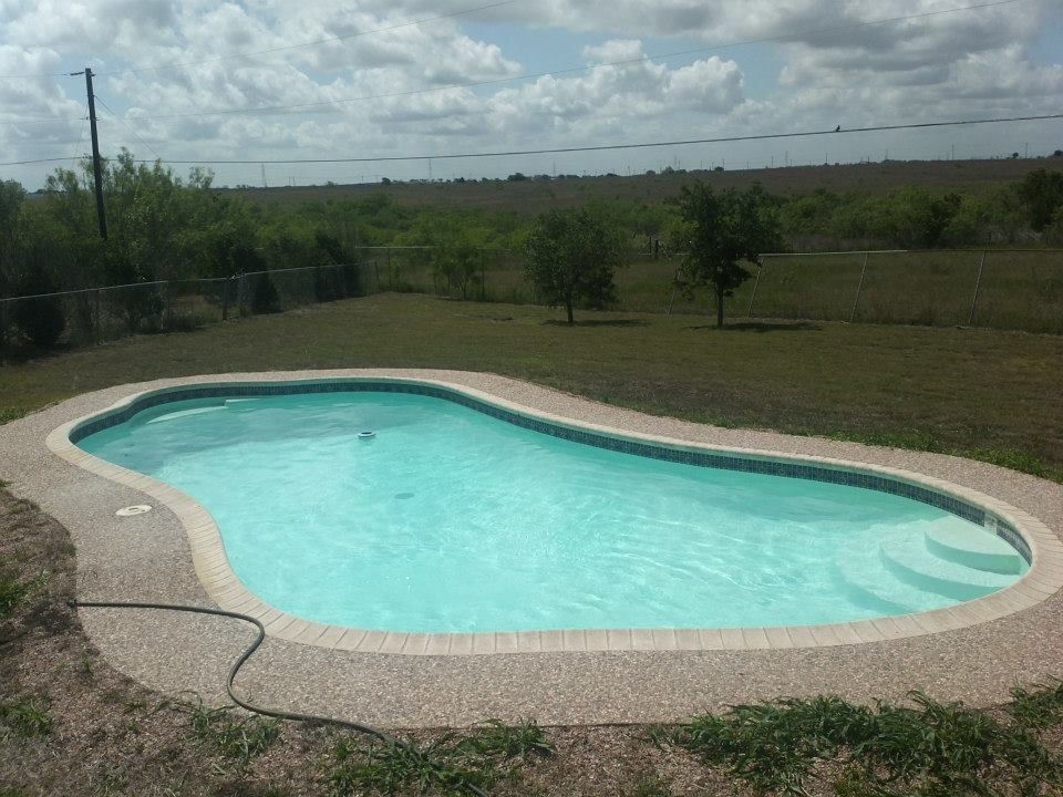 Outdoor kidney-shaped swimming pool with turquoise water, surrounded by gravel and a grassy yard under a cloudy sky