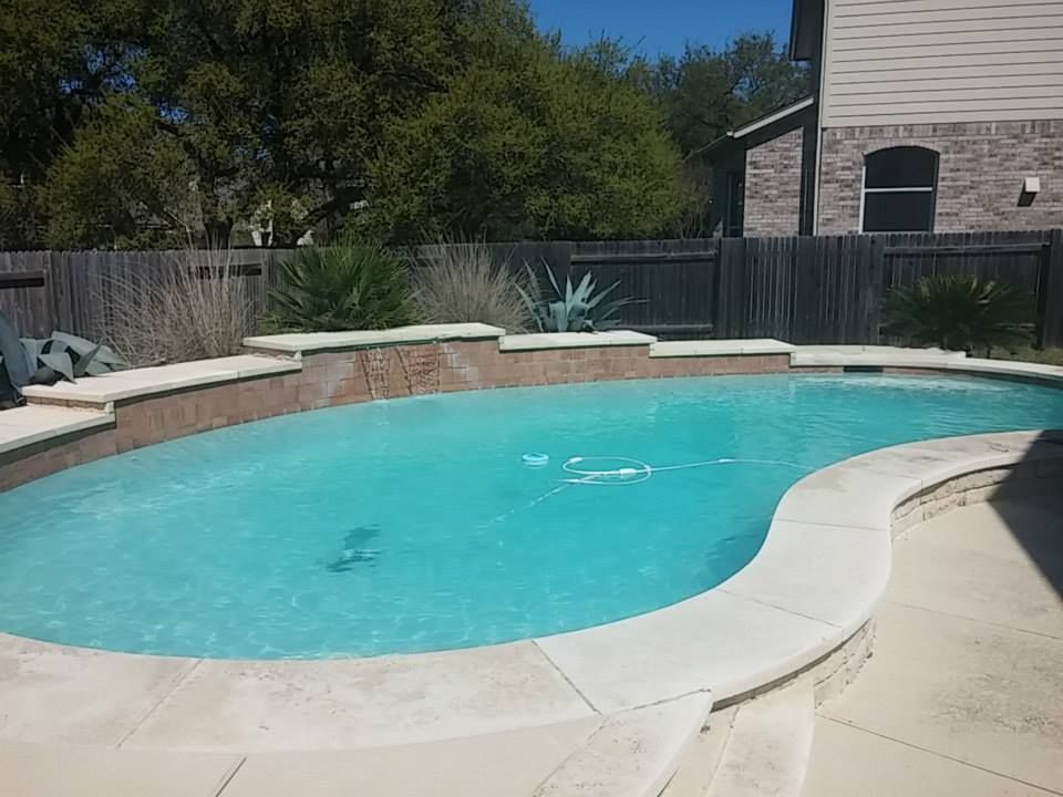 Curved backyard swimming pool with turquoise water, stone deck, fence, and trees in sunlight