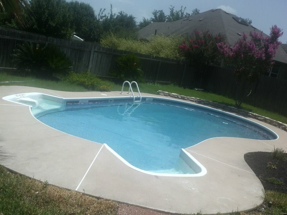 Backyard swimming pool with light blue water, white deck, and a metal ladder, surrounded by greenery