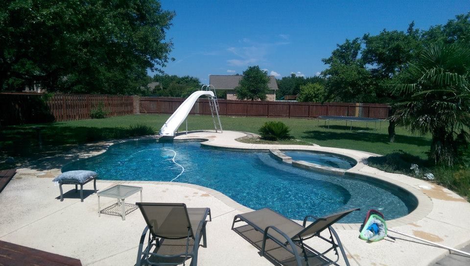 Backyard pool with a rock waterfall, lounge chairs, and a fenced lawn in bright sunlight