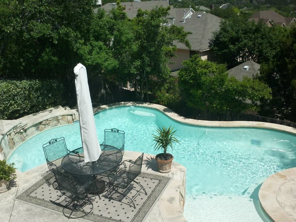Backyard pool with patio table, chairs, umbrella, and potted plant surrounded by greenery