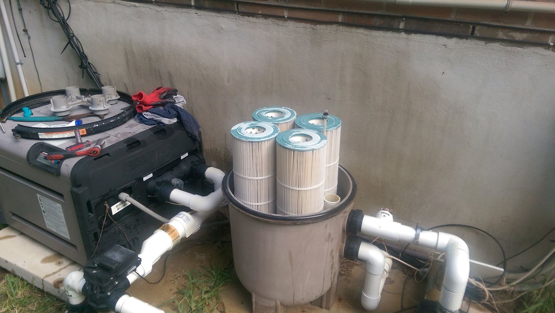 Pool filtration equipment with metal canister filter and pipes beside a concrete wall outdoors