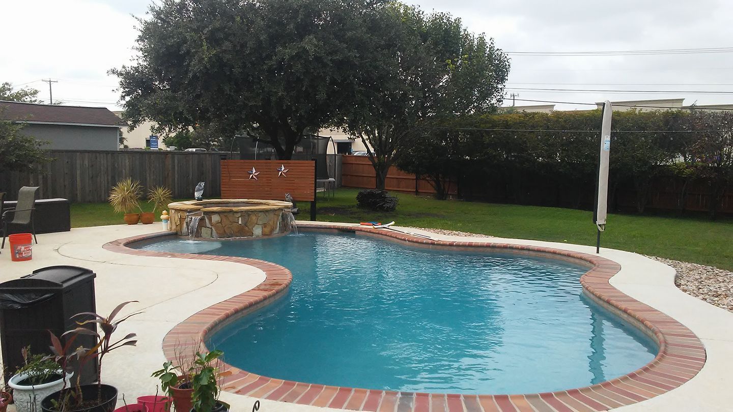 Backyard pool with curved stone edge, patio chairs, and large tree beside a brick wall