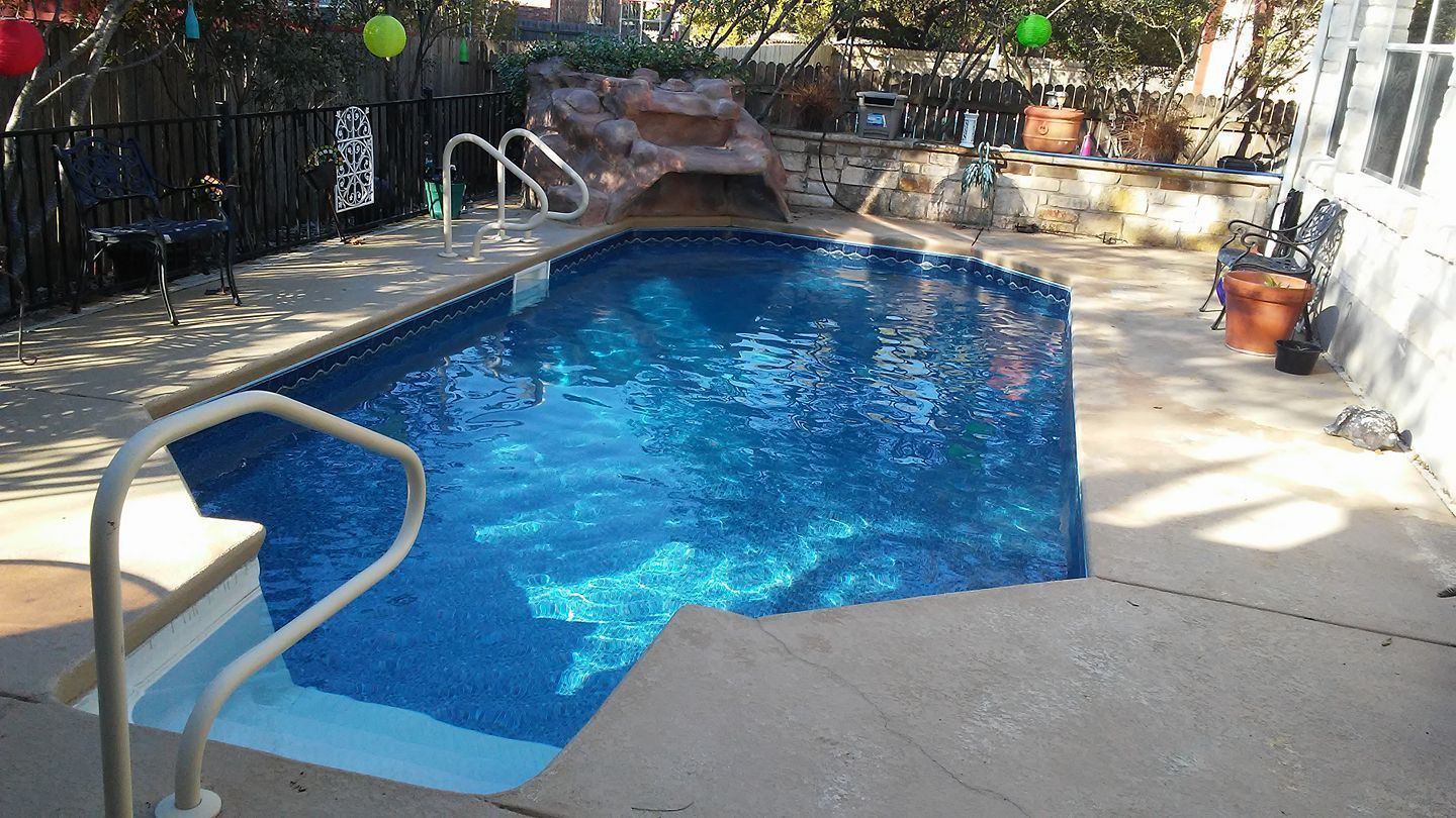 Backyard swimming pool with blue water, stone deck, and a metal handrail under bright sunlight