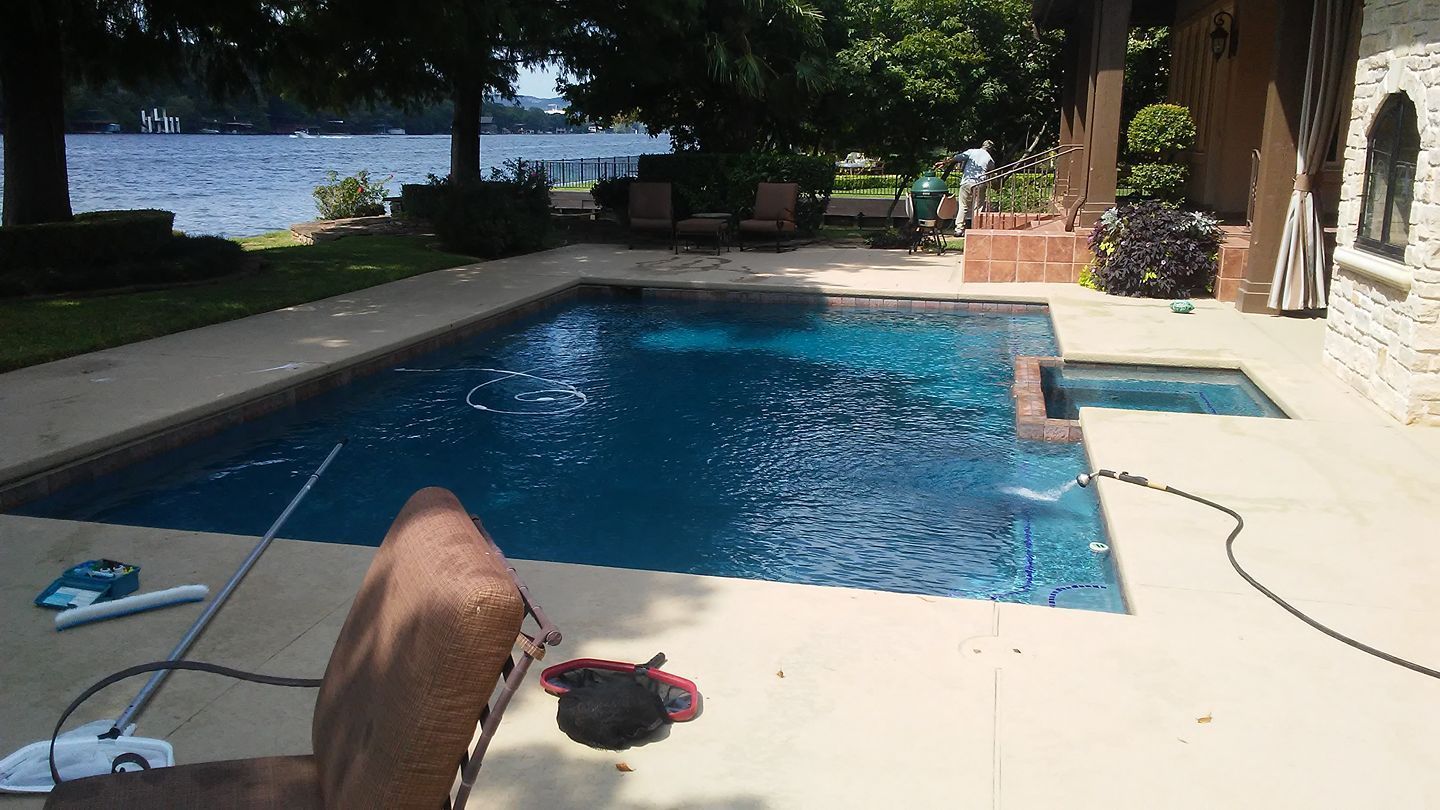 Backyard swimming pool beside a house, with lake view and patio furniture in the background.