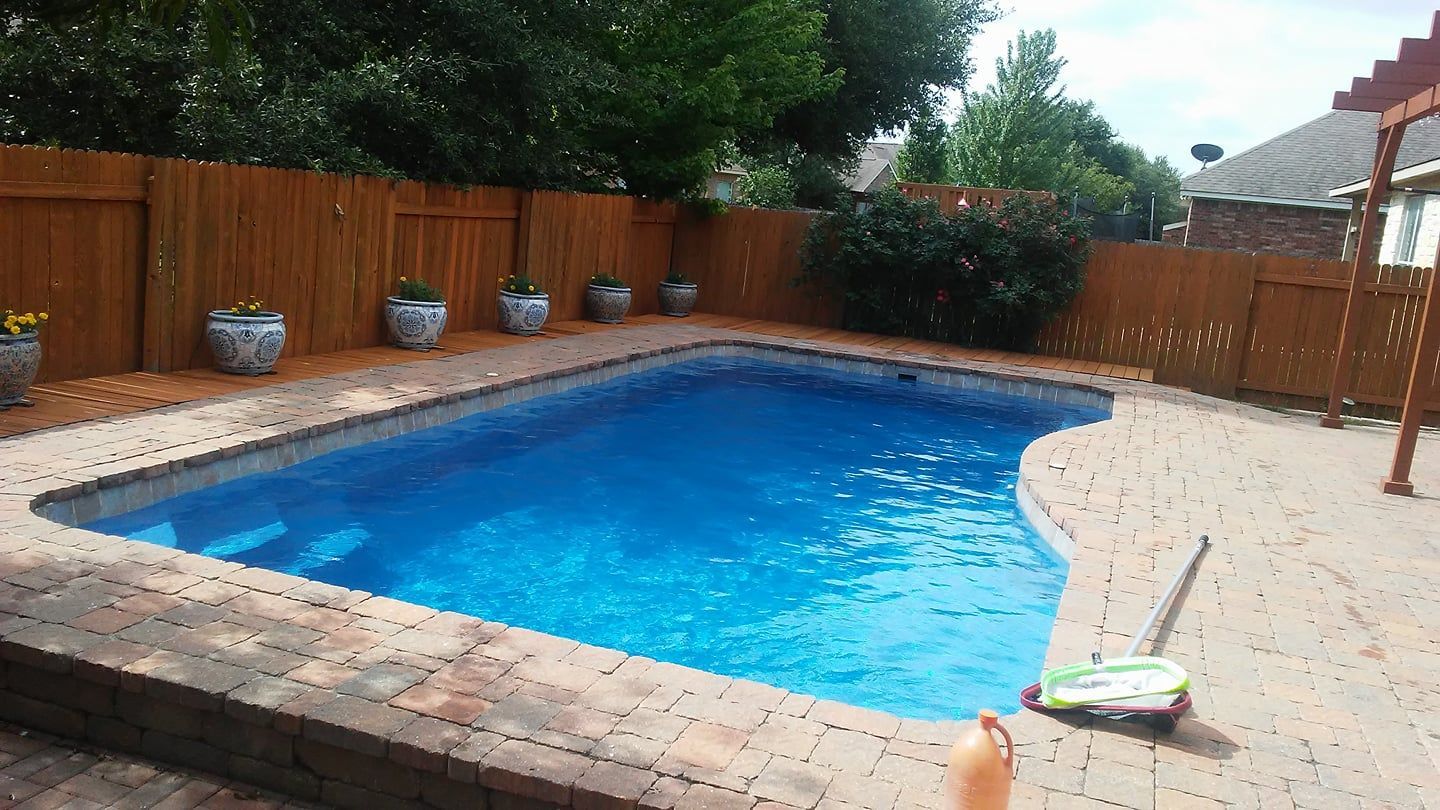 Backyard swimming pool with blue water, brick border, potted plants, and lounge chair on the deck