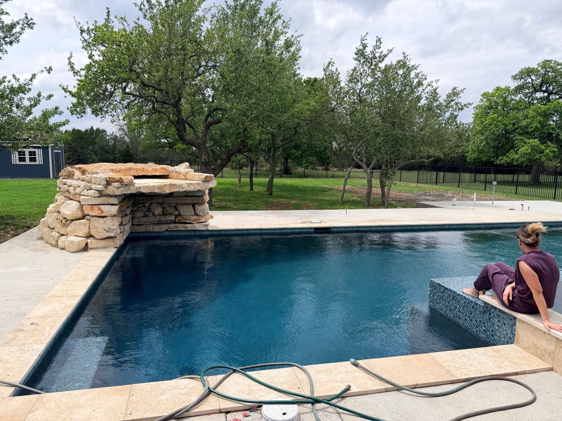 Backyard pool with a rock waterfall and a person sitting at the edge under trees