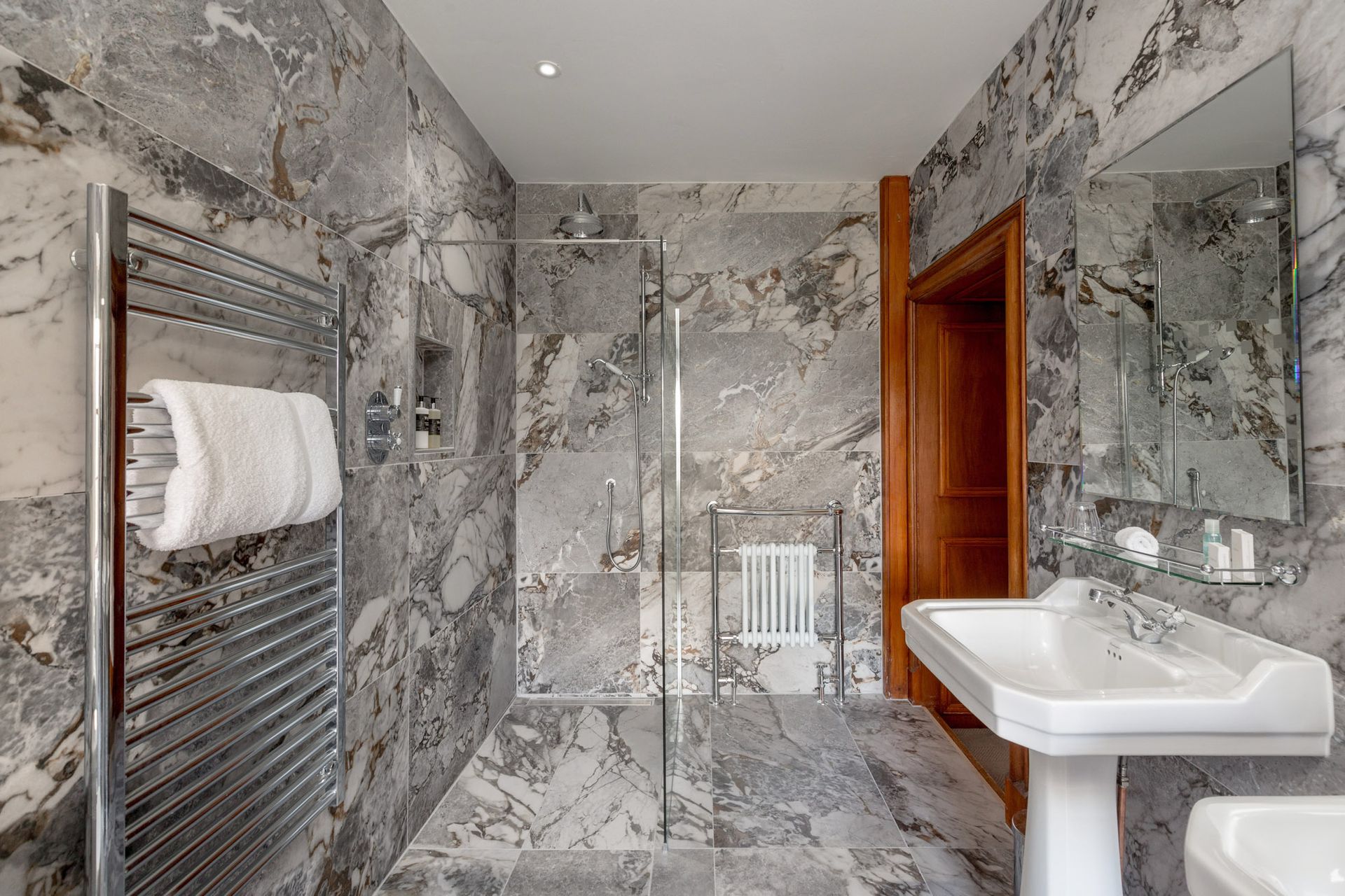 A bathroom with marble walls, a walk in shower, a sink and a mirror.