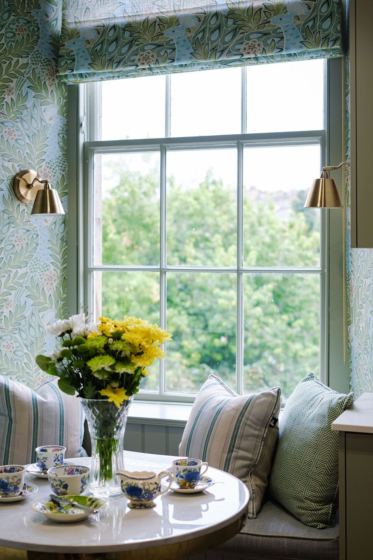 A cozy kitchen window seat with flowers, teacups, and pillows, overlooking greenery.
