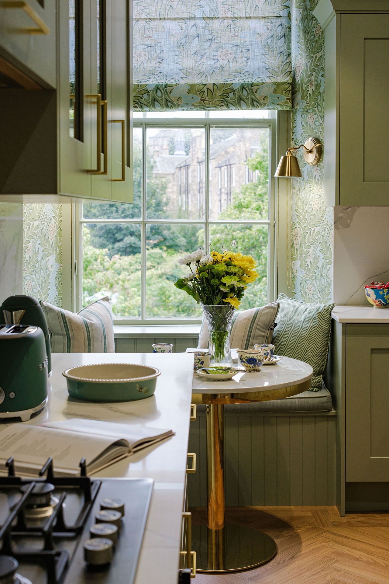 Kitchen nook with round table, window seat, and marble countertop; sage green cabinets and floral wallpaper.