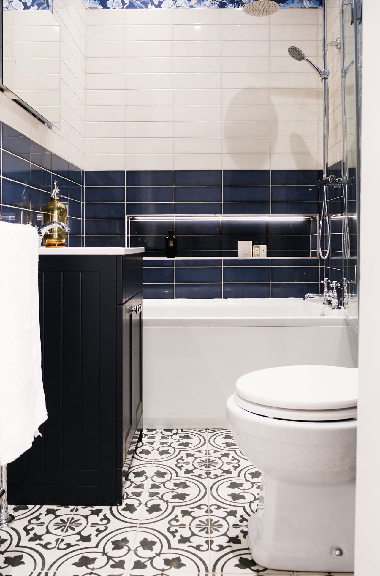 Bathroom with patterned floor tiles, white and blue tiled walls, dark blue cabinet, white toilet and bathtub.