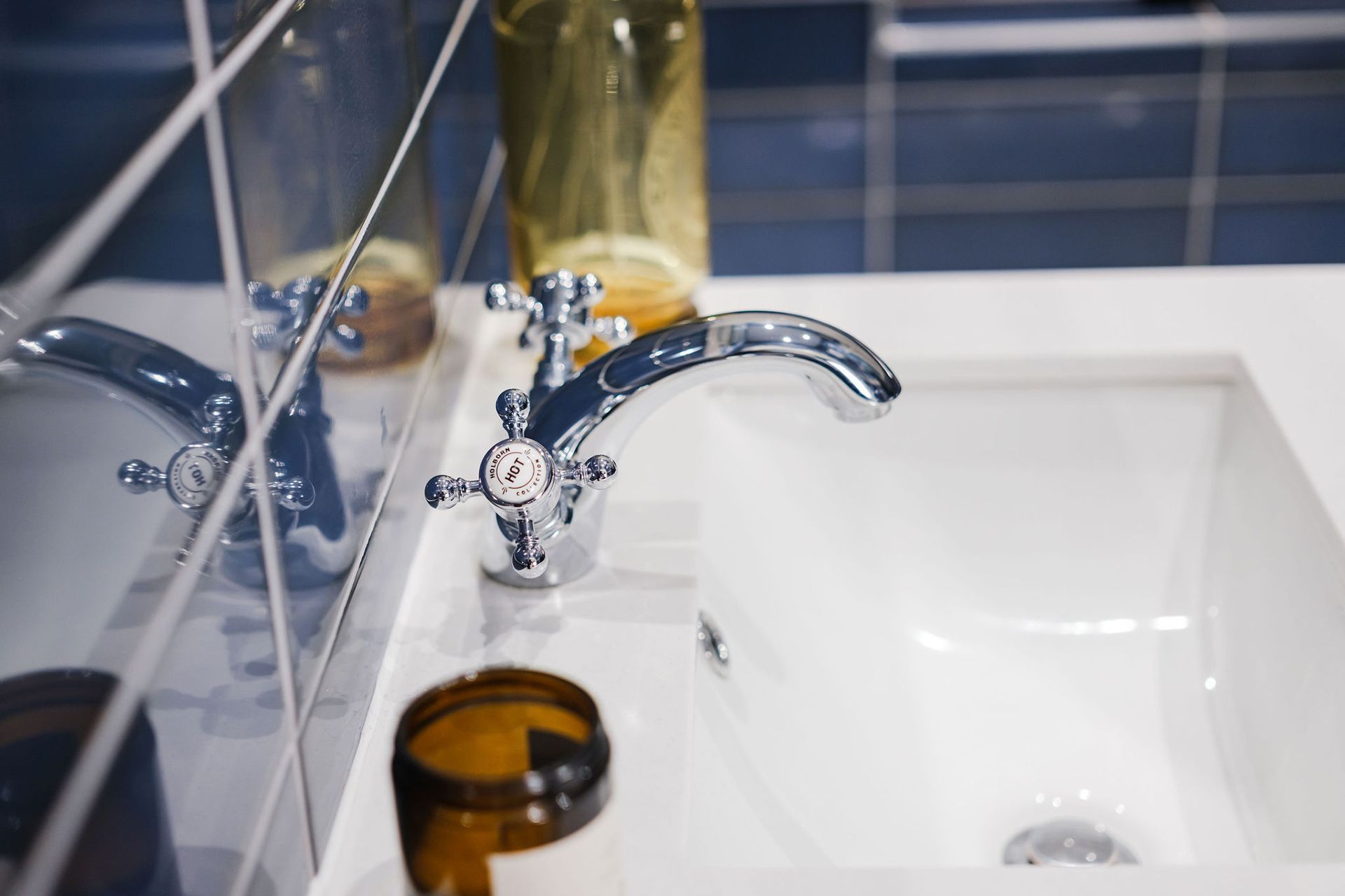 Close-up of a chrome faucet over a white sink, with blue tiled walls and bottles in the background.