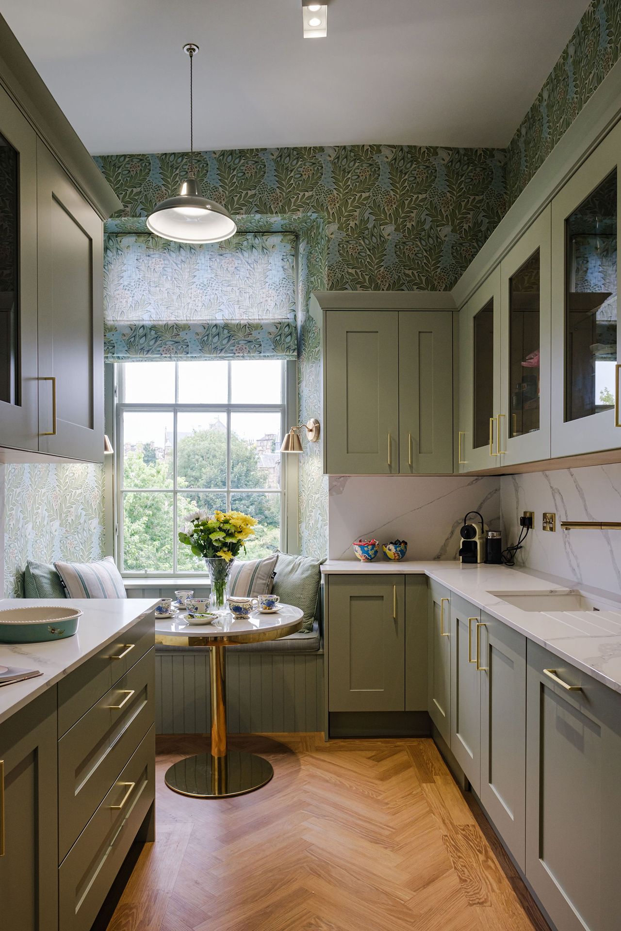 Small kitchen with green cabinets, floral wallpaper and built-in window seat with small table.
