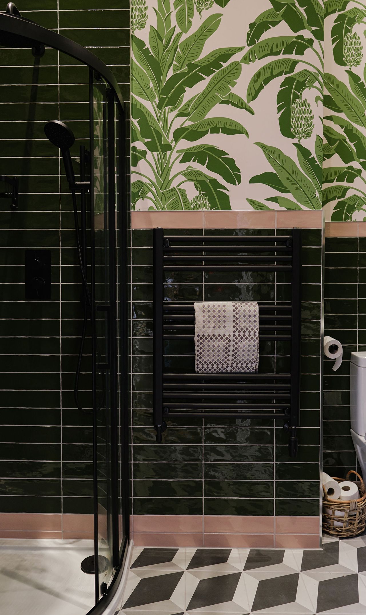 Bathroom with dark green tiled walls, tropical wallpaper, black towel rack and patterned floor.