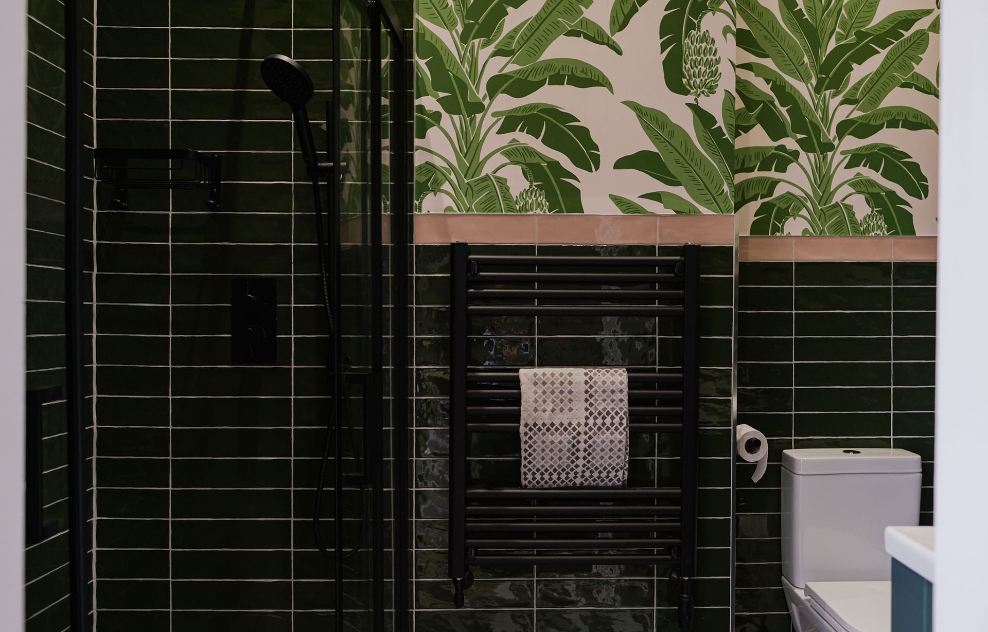 Bathroom with green tiled walls, black shower and tropical wallpaper. Black towel rack with towel. White toilet.