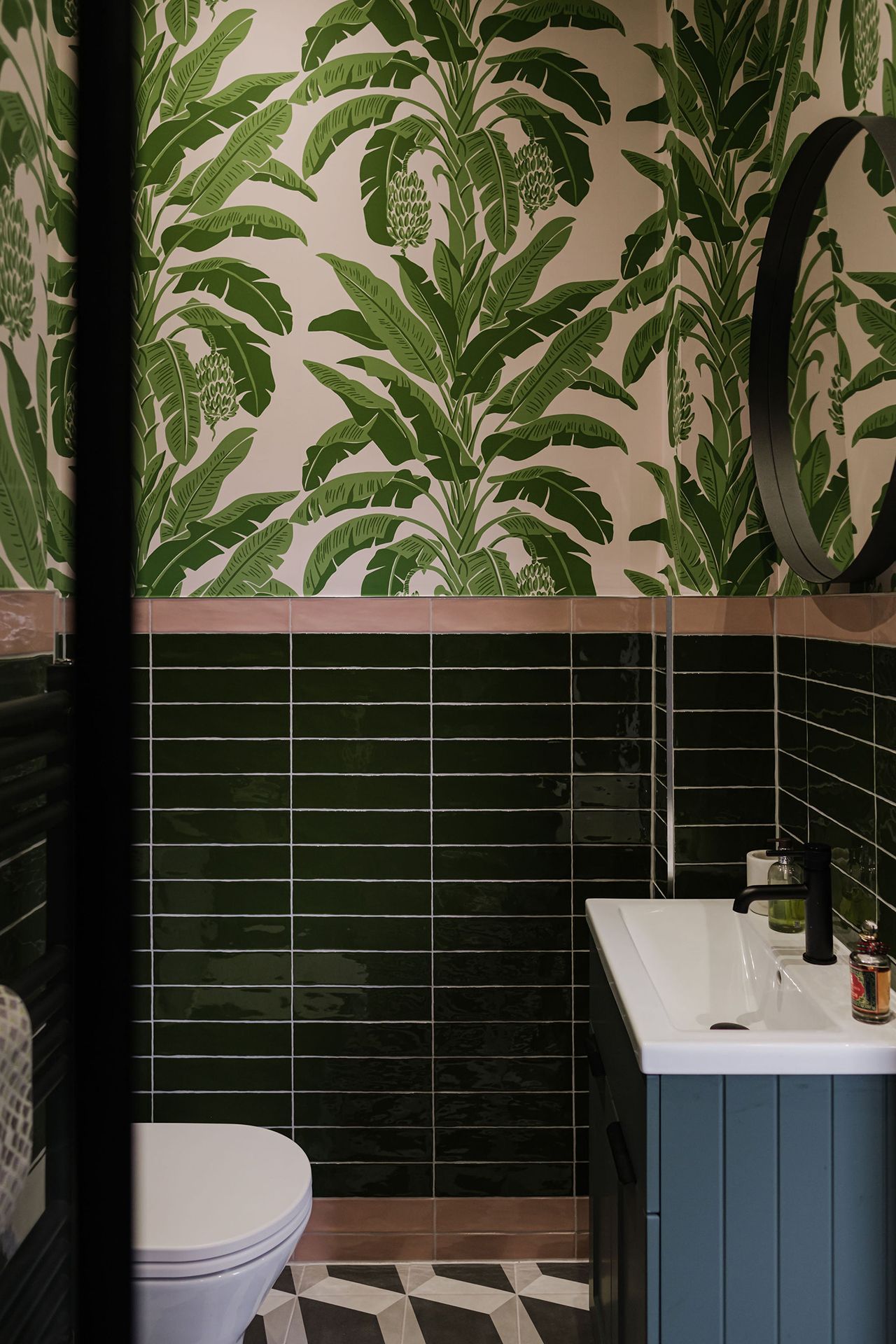 Bathroom with green botanical wallpaper, emerald tile, blue vanity and toilet.