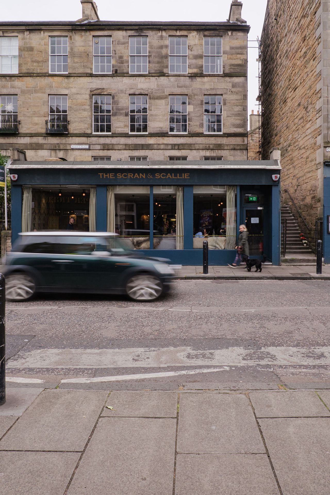 A green car speeds past a blue storefront on a cobblestone street; a person walks by with a cart.
