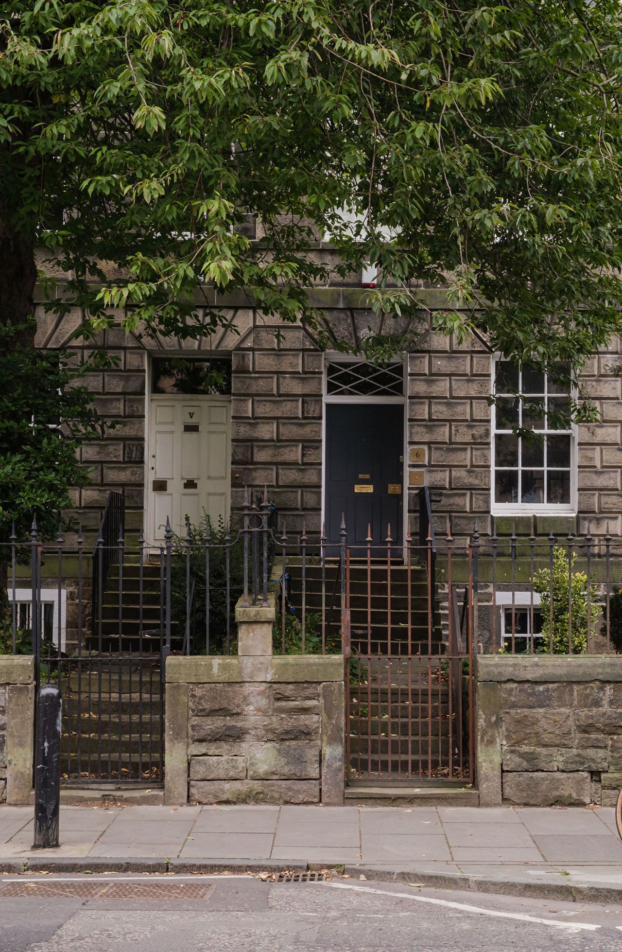 Stone buildings with doors, windows and wrought-iron fences under a leafy tree in an urban setting.
