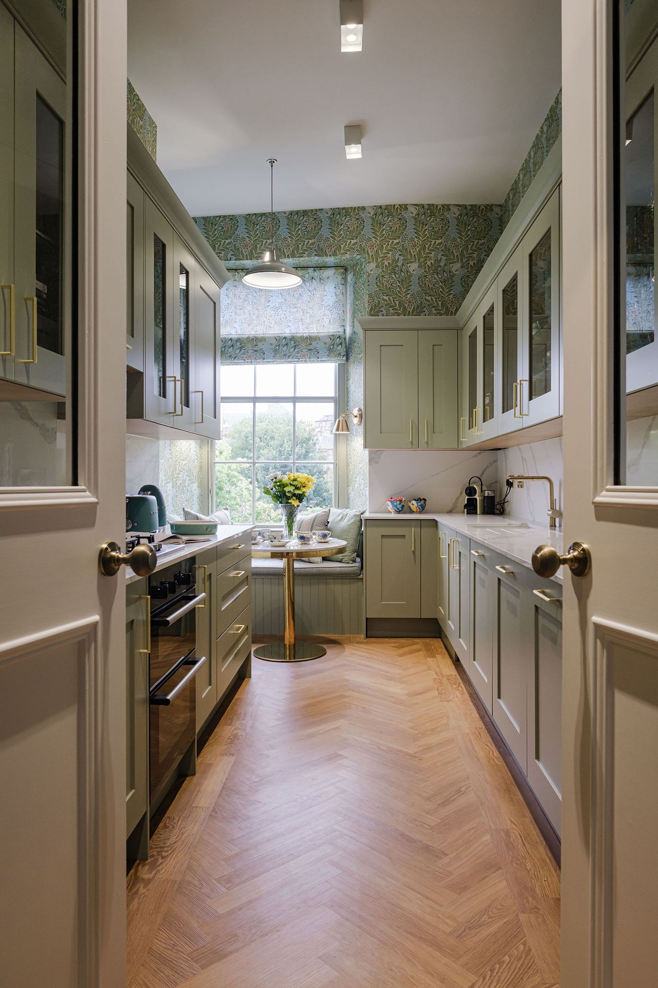Narrow, light green kitchen with floral wallpaper, a window seat and a herringbone floor.