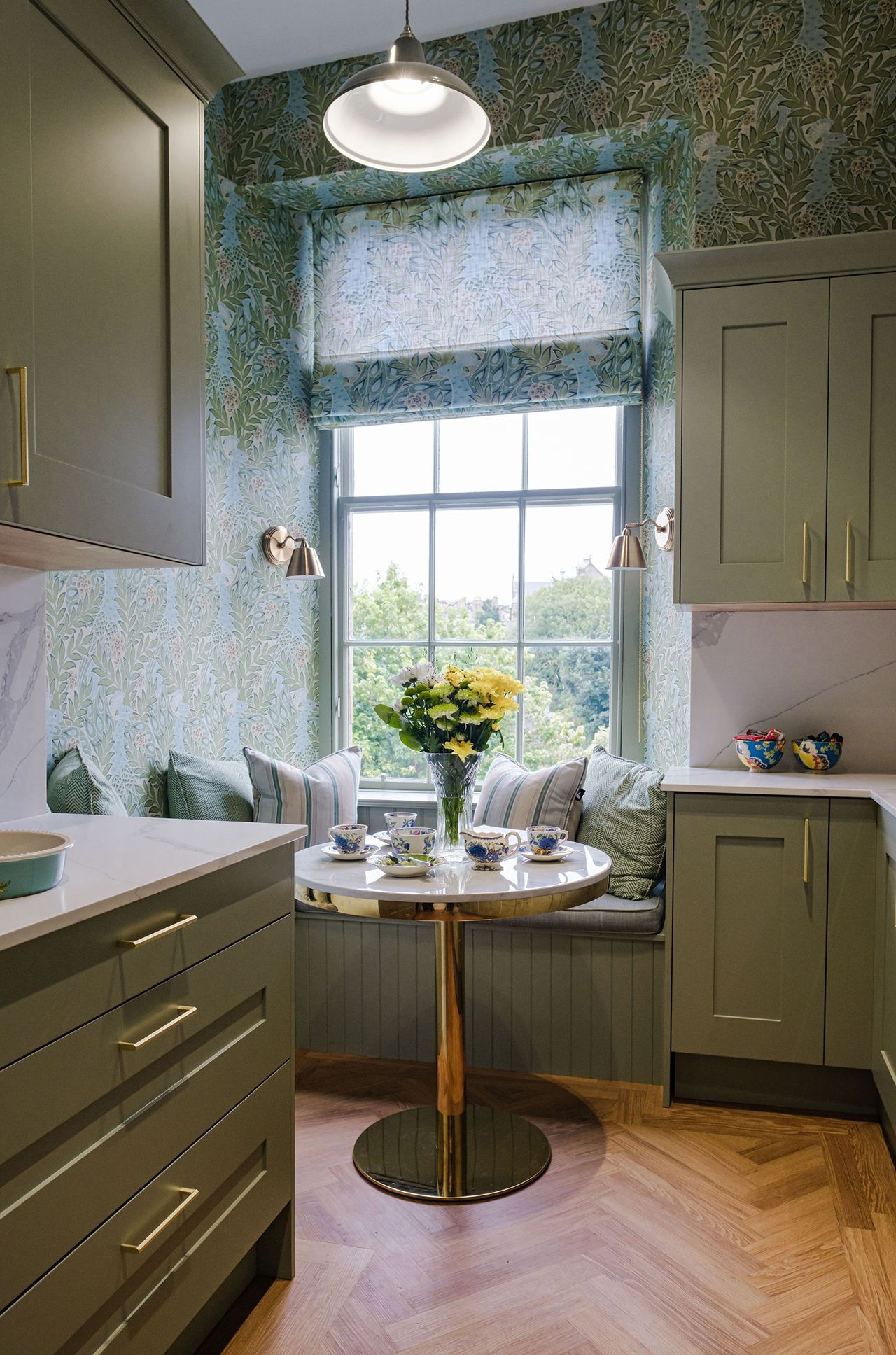 Green kitchen with window seat, floral wallpaper, round table set for tea.