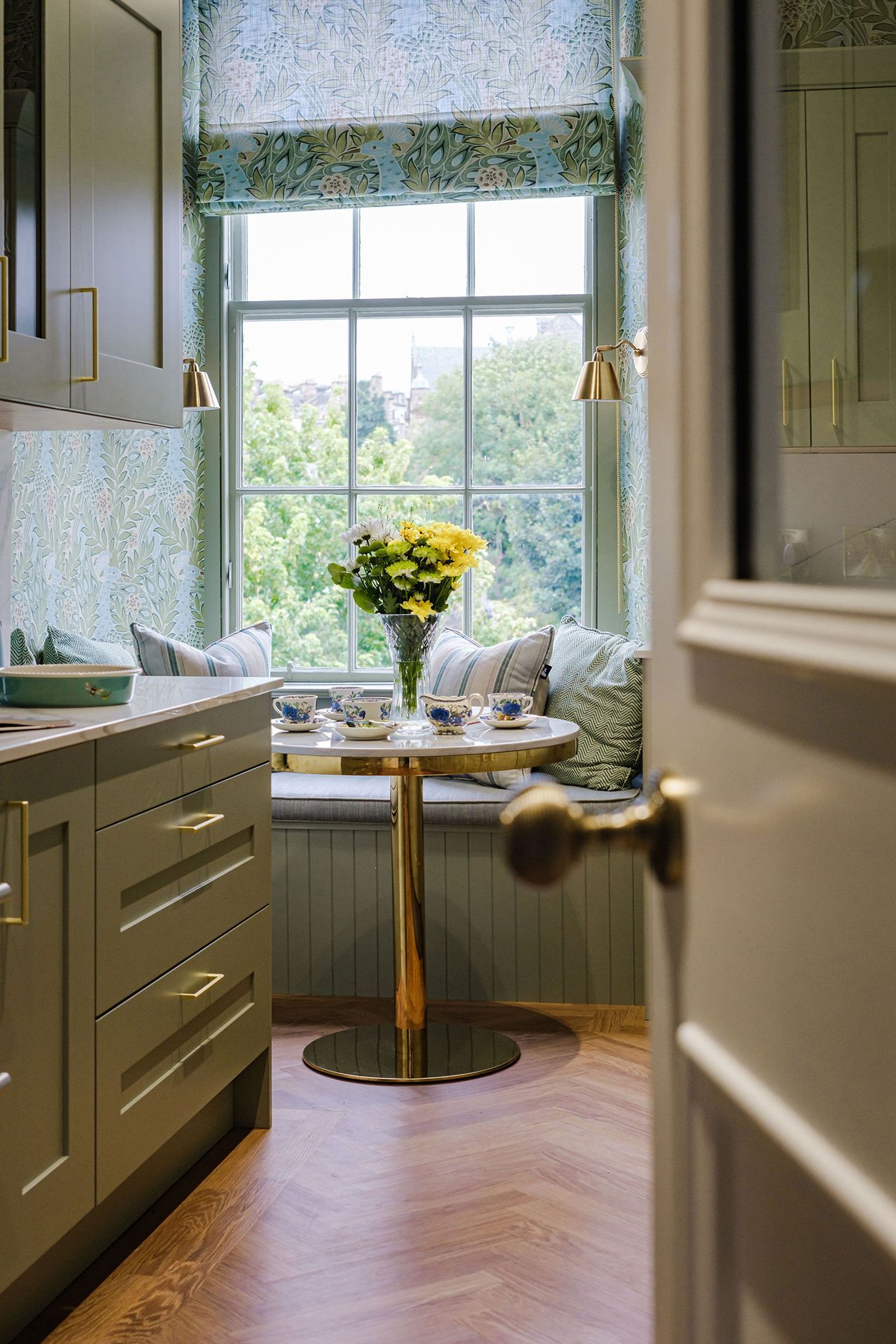 Kitchen with window seat, floral wallpaper, round table with flowers, green cabinetry and door handle in foreground.