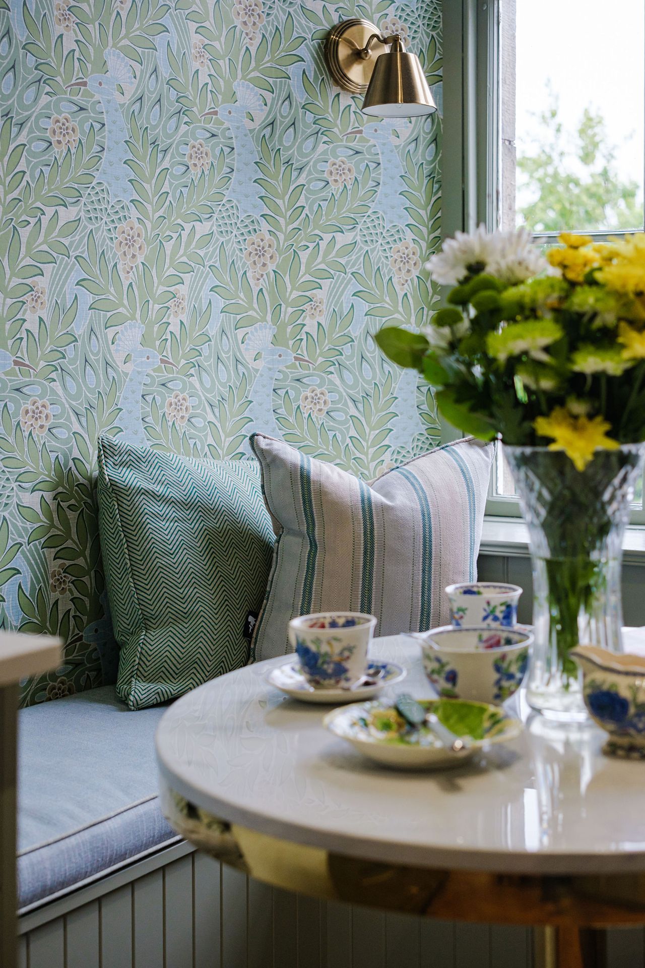 Cozy nook with floral wallpaper, cushioned bench, pillows, and a table set with teacups, illuminated by a wall sconce.