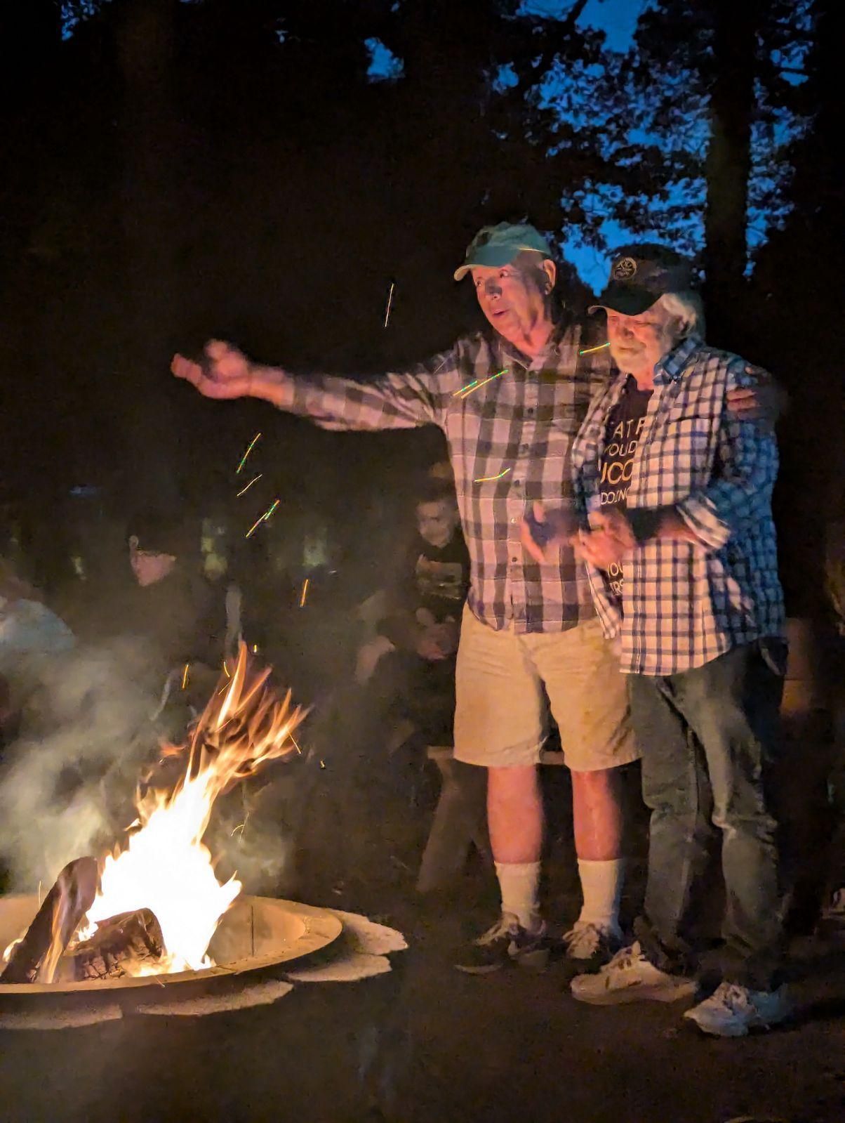 Two men are standing next to a fire pit at night.