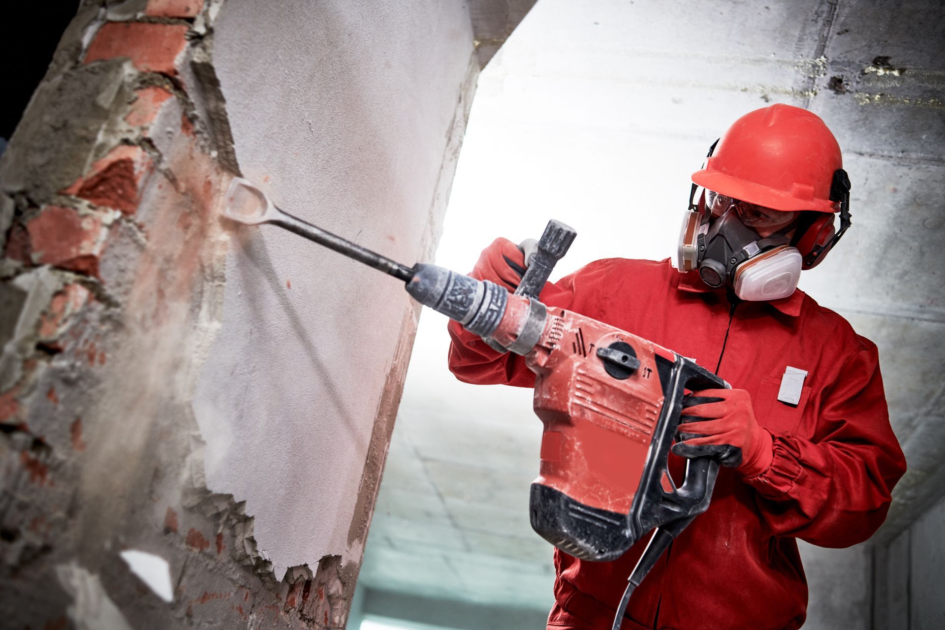 A demolition worker wearing a face mask uses a hammer to break an interior wall's plastering.