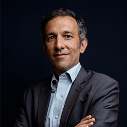 Man with dark hair in suit, arms crossed, smiling against a dark background.